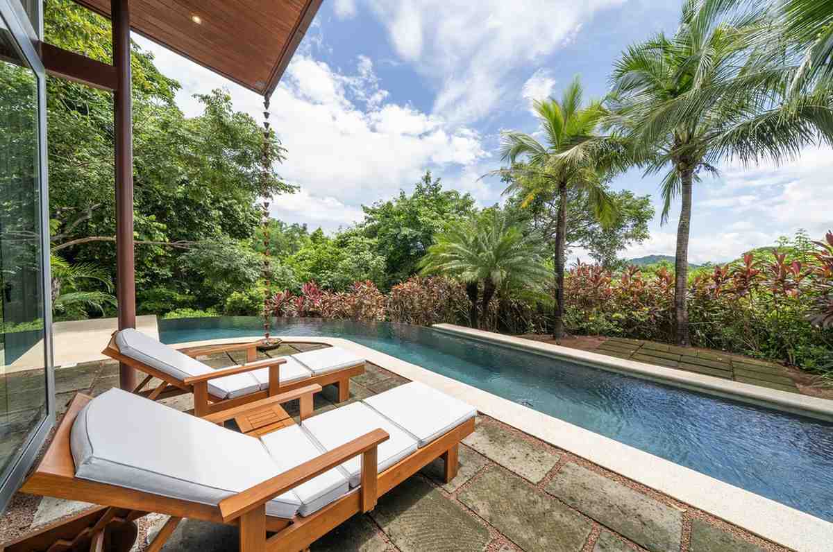 Wooden lounge chairs on a stone deck positioned next to a sleek swimming pool bordering lush landscaping.