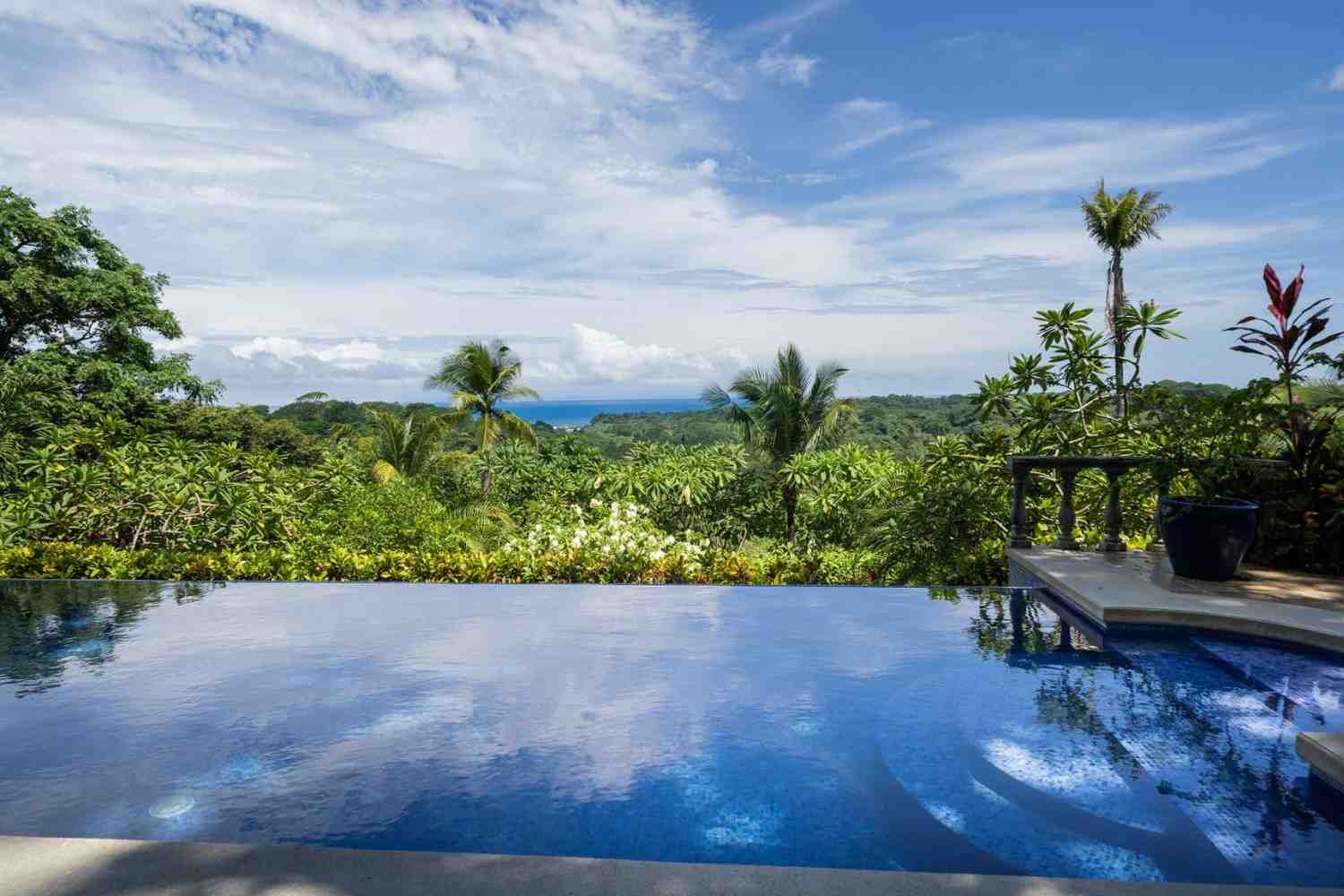 Edge of an infinity pool reflecting the sky, overlooking lush green treetops and distant ocean waters.