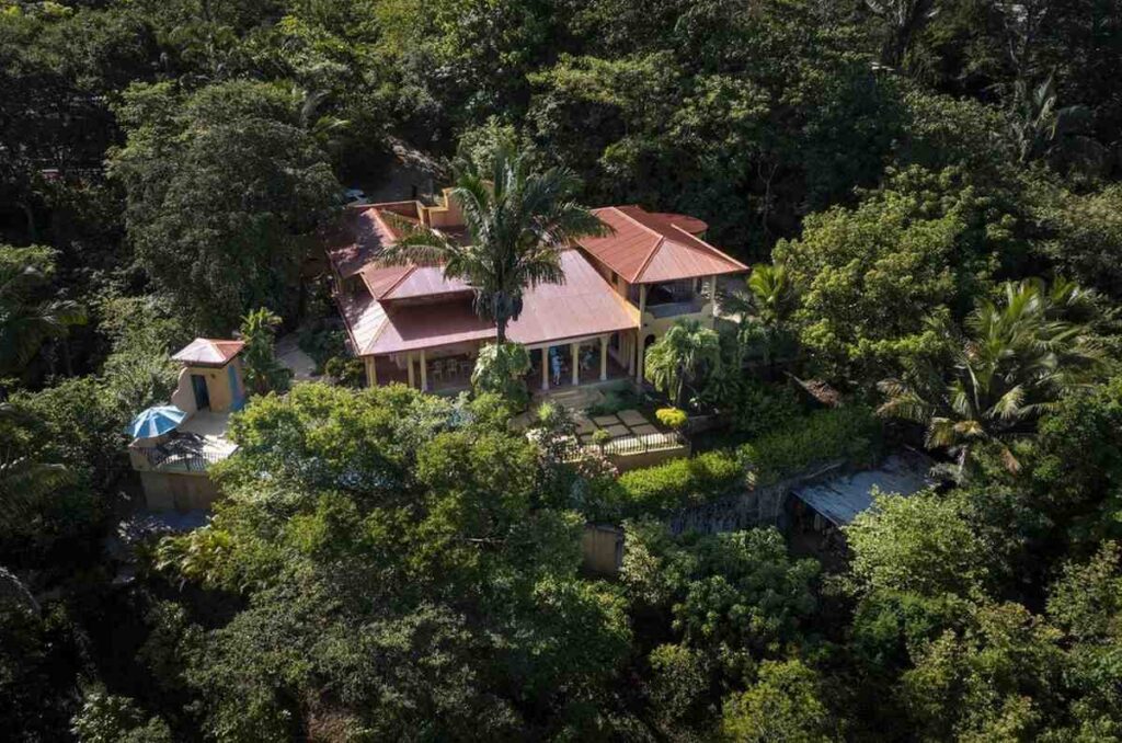 Angled aerial perspective highlighting the multi-tiered red roof structure of the colonial house surrounded by thick forest foliage.