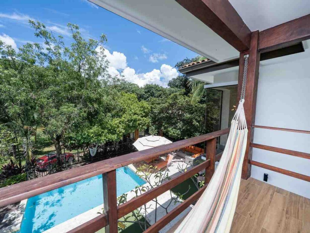 Angle from a covered wooden balcony with a hammock looking down toward a blue swimming pool surrounded by tropical foliage.